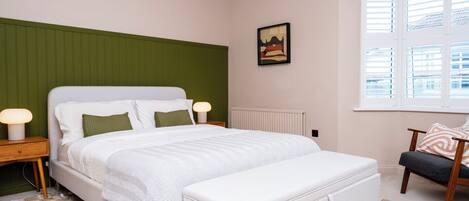 A bedroom with a bed, bedside lamps, a radiator, a window with blinds, and a chair.