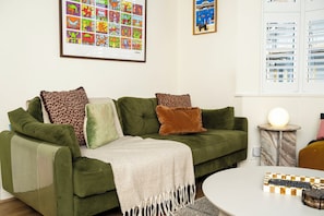 A green sofa with multiple cushions, a white throw blanket, a framed artwork above, and a round table with a remote control and a decorative box. - 2BDR Brighton Retreat with Courtyard Garden (Brighton and Hove)