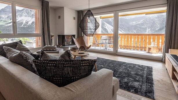 Ein modernes Wohnzimmer mit Sofa, Couchtisch und Fernseher. Große Fenster bieten Blick auf die Berge.