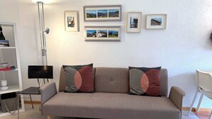 En soffa i en neutral färgskala med mönstrade kuddar, en golvlampa, ett litet bord med en lampa och en vägg med inramade tavlor.
