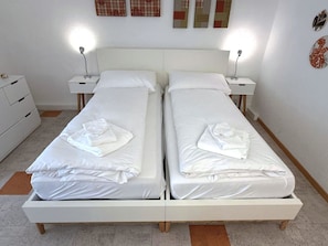 A double bed with white linens and a folded towel, two bedside tables with lamps, and a white dresser. - Fortuna 220 # 2-bed apartment, Dusche, WC 2-Bettwohnung, Dusche, WC by Interhome (Leukerbad)