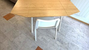 Ett matbord med en träskiva och vita stolar i ett rum med ett grått kaklat golv och en grå vägg.
