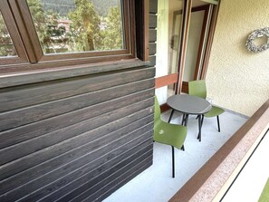 Un balcón con piso de madera, una pequeña mesa, dos sillas y vistas a un paisaje montañoso a través de una ventana.