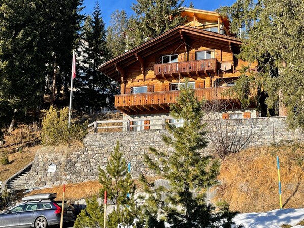Un chalet in legno a più piani con balconi, pareti in pietra e un'area di parcheggio con auto.