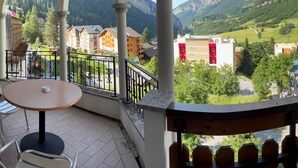 Ein Balkon mit Tisch und Stühlen, der auf eine gebirgige Landschaft mit Gebäuden und Grünblicken blickt.
