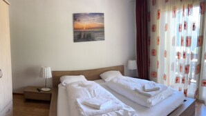 Ein Doppelbett mit weißer Bettwäsche und zwei Kissen, ein Nachttisch mit Lampe sowie ein Sonnenuntergangsbild an der Wand.