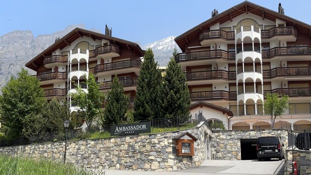 Ein Hotel mit mehreren Balkonen, einem Eingang mit Steinmauer und einem Berg im Hintergrund.