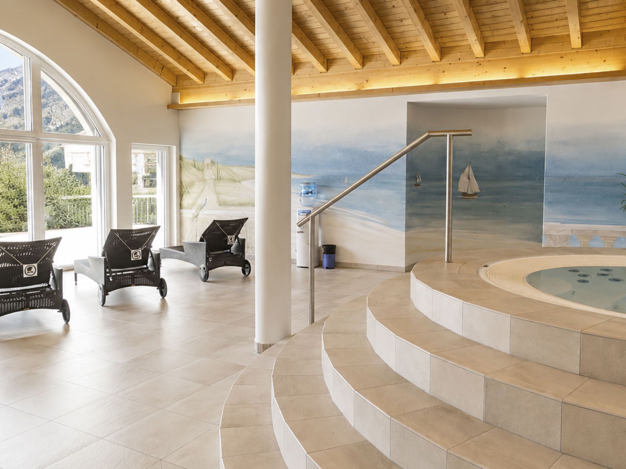 Ein großzügiger Innenbereich mit einer geschwungenen Treppe zu einem Whirlpool, großen Fenstern und einem Wandbild mit Strandmotiv.