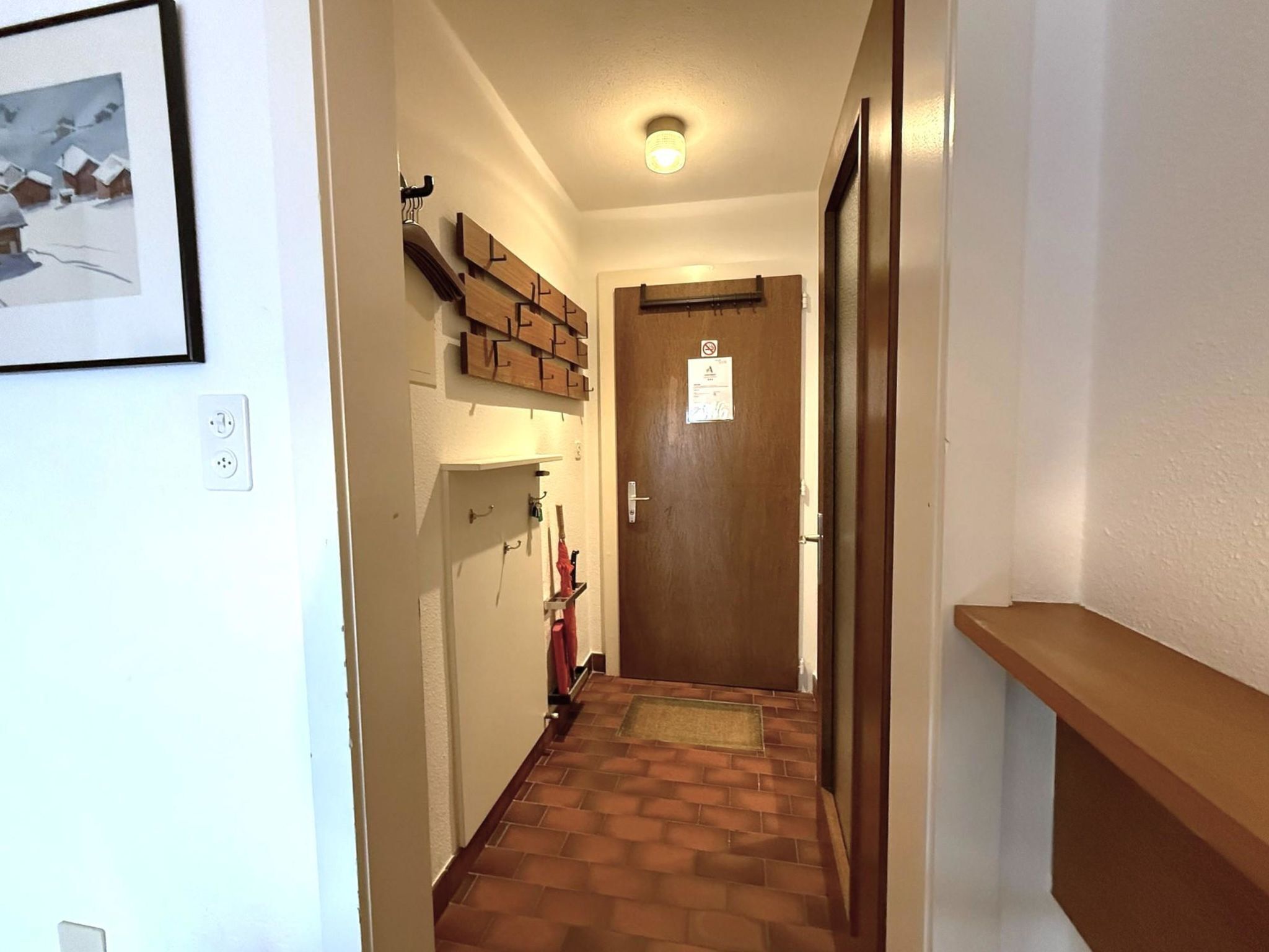 A narrow hallway with a wooden door at the end, a coat rack with items, and a framed picture on the wall.