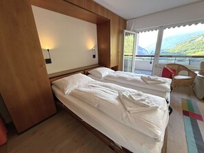 Hotelkamer met twee bedden, een houten hoofdbord, een balkon met uitzicht op de bergen en een rieten stoel.