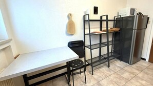 Kompakti keittiö, jossa on valkoinen työtaso, musta tuoli, puinen leikkuulauta, musta metallinen hyllykkö ja ruostumattomasta teräksestä valmistettu jääkaappi.