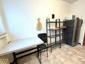 A compact kitchen with a white countertop, a black chair, a wooden cutting board, a black metal shelf unit, and a stainless steel refrigerator. - Salve 9 # 6-bed apartment, Dusche, WC 6-Bettwohnung, Dusche, WC by Interhome (Leukerbad)