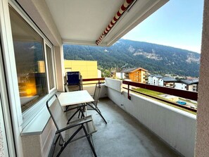 A balcony with a table and chairs, a view of a mountain, and a yellow building in the background. - Salve 9 # 6-bed apartment, Dusche, WC 6-Bettwohnung, Dusche, WC by Interhome (Leukerbad)