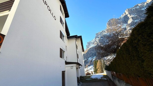 Eine schmale Straße mit weißen Gebäuden, eines mit der Aufschrift 'APOLLO', die zu einer Bergblick führt.