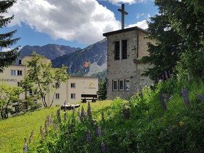 A hotel with a mountain backdrop, a chapel with a cross, and a signboard. - Chalet Willi-1.OG Appartement/Fewo, Dusche und Bad, WC, 9-Bettwohnung by Interhome (Arosa)