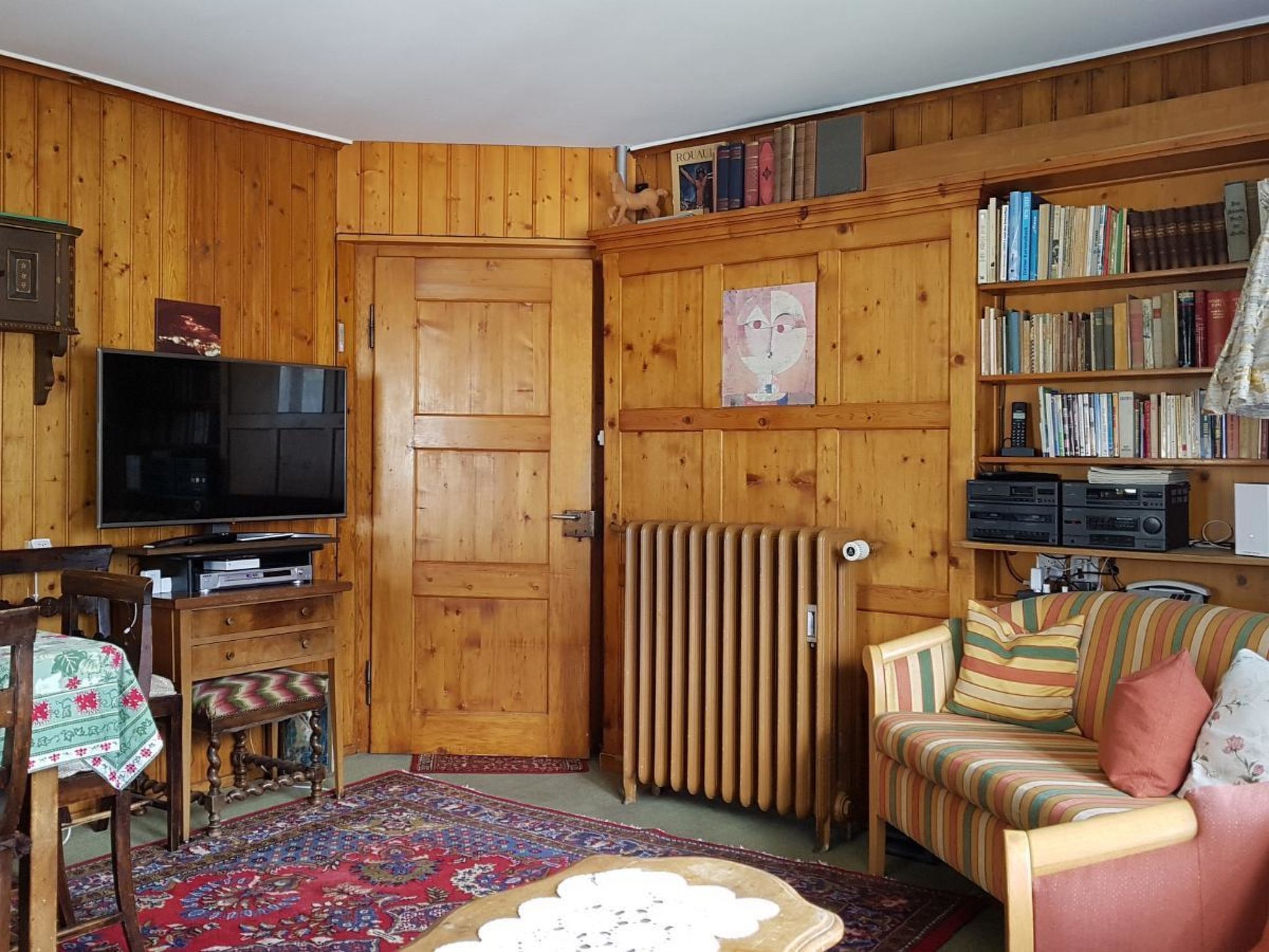 A cozy living room with wooden walls, a television, a bookshelf, a radiator, and a striped sofa.