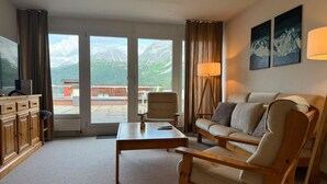 Ein Wohnzimmer mit einer Couch, einem Sessel, einem Couchtisch und einem Fernseher auf einem Schrank. Große Fenster bieten Blick auf die Berge.