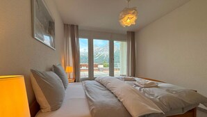 Ein Hotelzimmer mit Bett, Nachttisch, Lampe, Blick auf Berge durch das Fenster und einer Deckenleuchte.