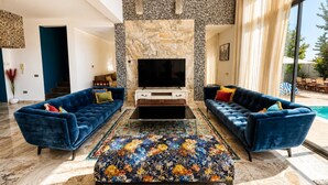 Un salón amplio con un televisor de pantalla plana grande, dos sofás azules, un reposapiés estampado y una pared de acento de piedra.