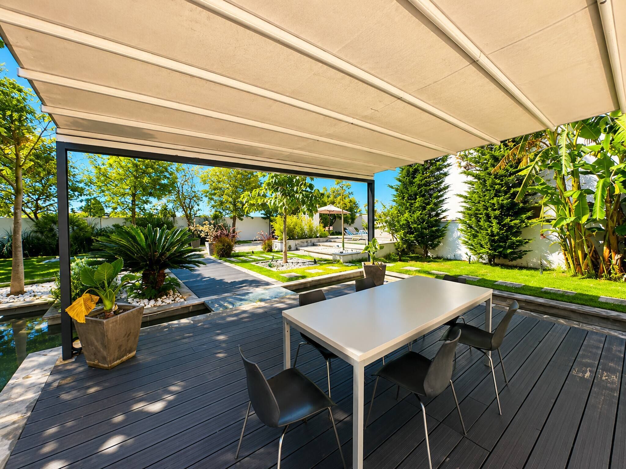 A modern outdoor patio with a table and chairs, surrounded by greenery and a view of a swimming pool.