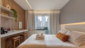 A modern hotel room with a bed, a small kitchenette, a window with curtains, and a view of buildings. - Film studio with parking space - 800 m from the park. Ibirapuera (São Paulo)
