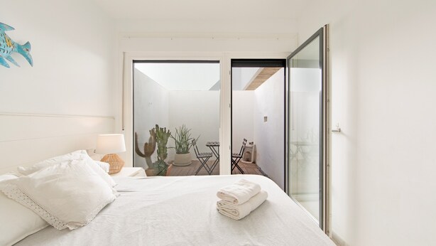 A bedroom with a bed, a nightstand, a lamp, a cactus, a chair, and a view of an outdoor area.