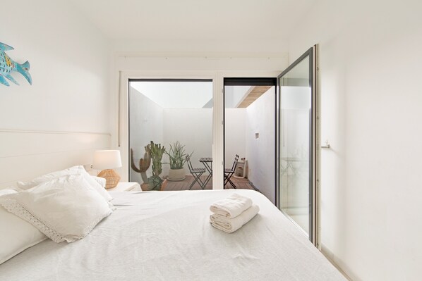 Un dormitorio con cama, mesita de noche, lámpara, un cactus, una silla y vistas a un espacio exterior.