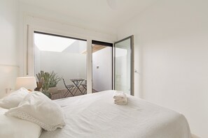 A bedroom with a bed, a nightstand, a lamp, a potted plant, and a view of an outdoor patio with a table and chairs.