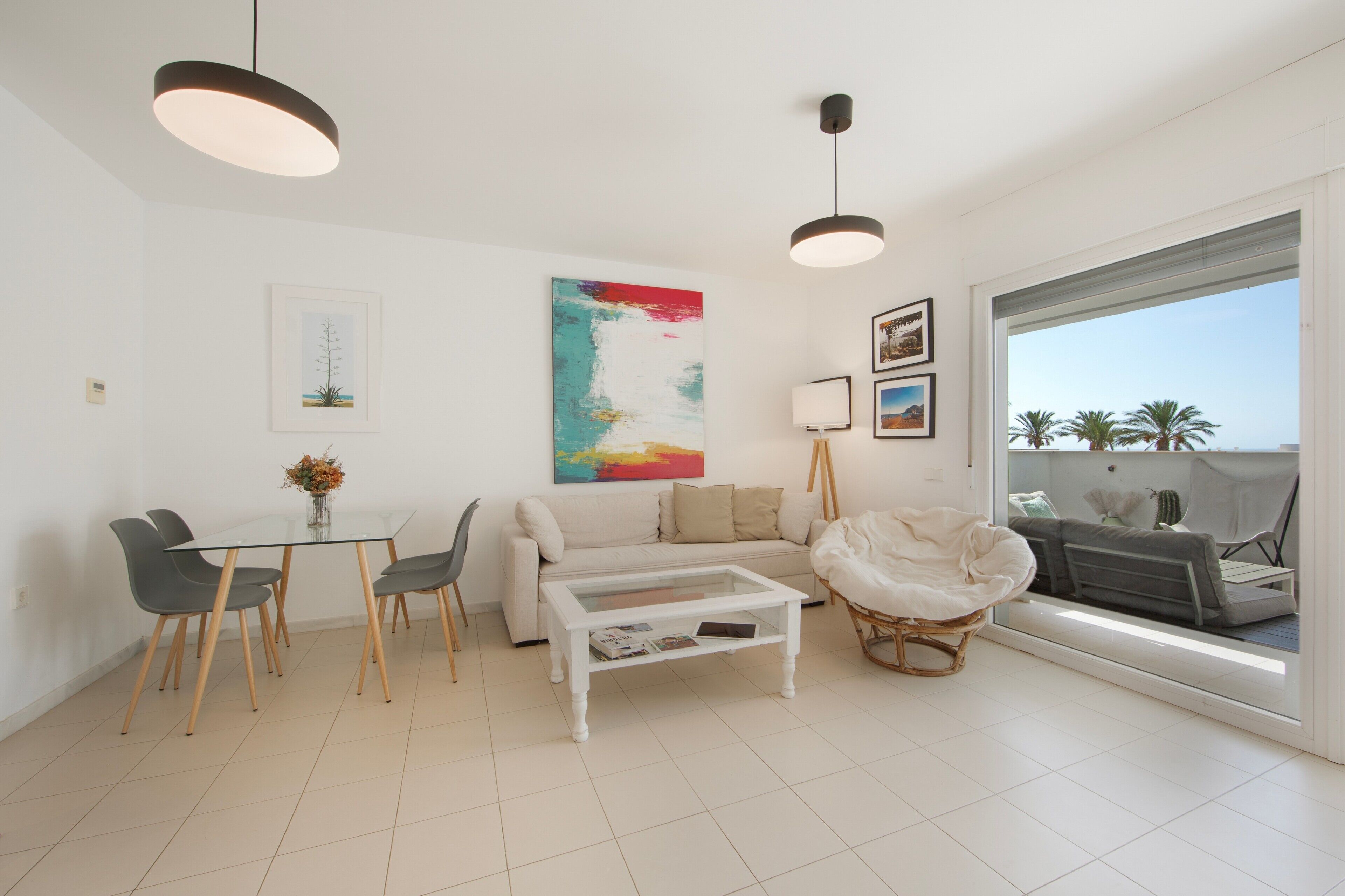 A modern living room with a glass dining table, a sofa, a coffee table, and a large window offering a view of palm trees.
