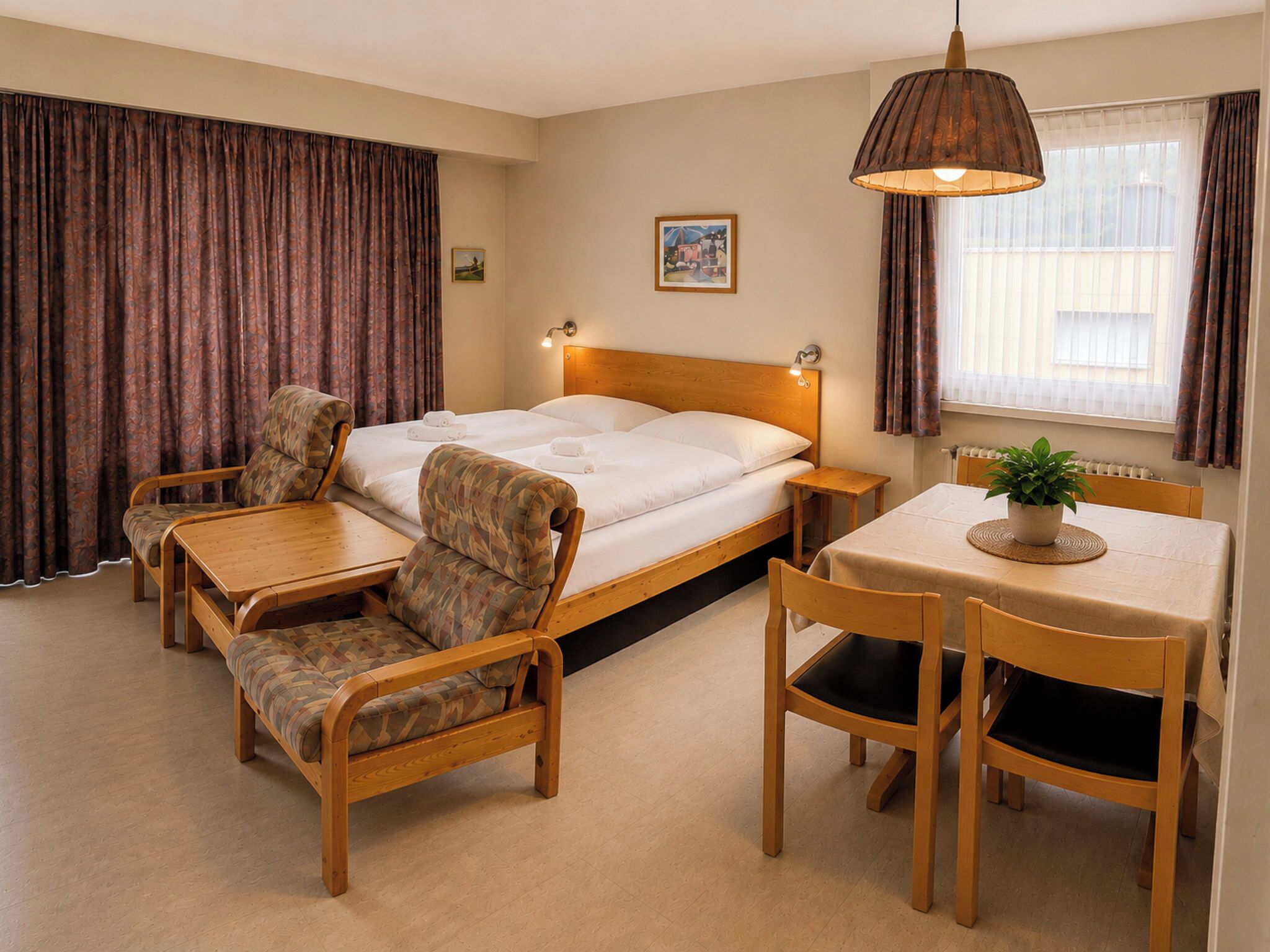 Une chambre d’hôtel comprenant un lit, une petite table avec des chaises, un canapé, une fenêtre avec des rideaux et une lampe fixée au mur.
