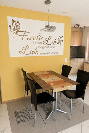 A dining area with a table and chairs, a kitchen in the background, and a wall with a decorative sign. - Majesta Leukerbad # 4-bed apartment, Dusche, WC by Interhome (Leukerbad)