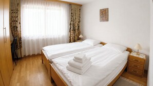 Hotellihuone, jossa on kaksi sänkyä, puulattia, työpöytä lampun kanssa ja ikkuna, jossa on ohuet verhot.