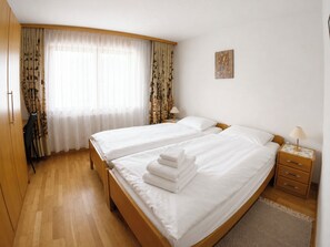 Ein Hotelzimmer mit zwei Betten, Holzboden, Schreibtisch mit Lampe und einem Fenster mit durchsichtigen Vorhängen.