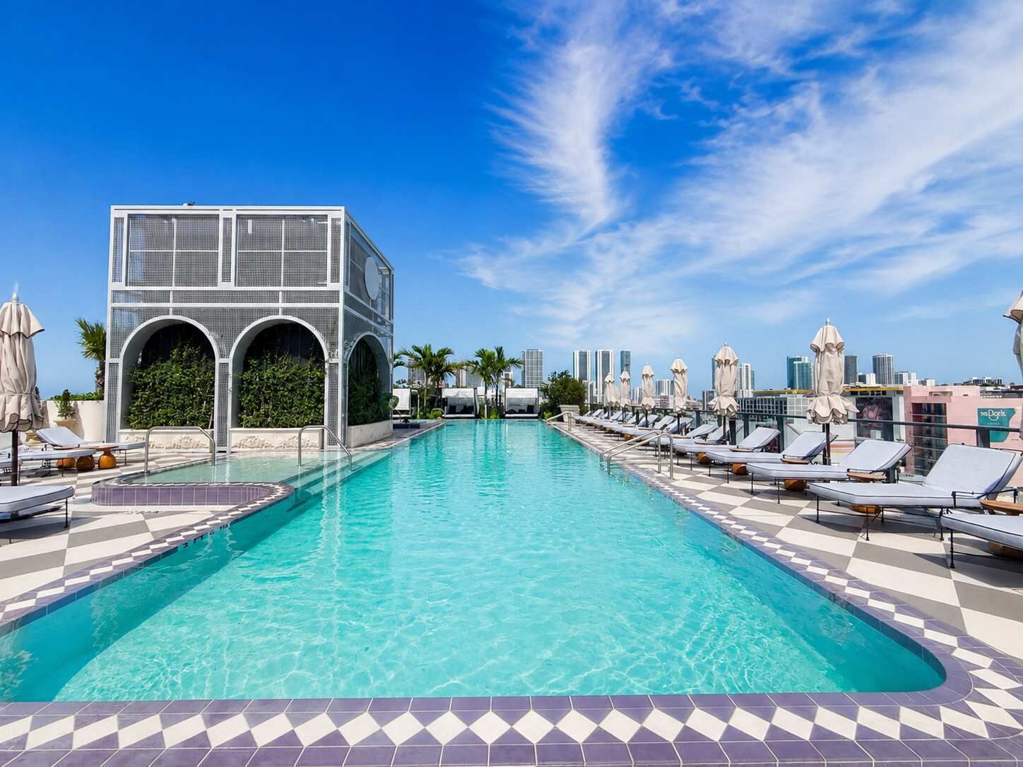 Uma piscina com espreguiçadeiras e guarda-sóis, um edifício moderno com janelas em arco e um horizonte urbano ao fundo.