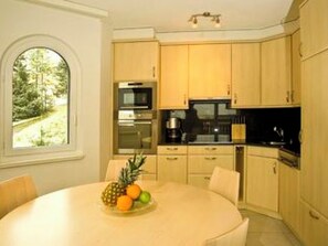 A modern kitchen with wooden cabinets, a round dining table with fruit, and a view of a forest through a window. - Ambassador A11 # 4-bed apartment, Dusche und Bad, WC by Interhome (Leukerbad)
