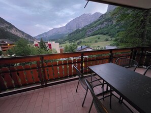 Un balcon avec une table et des chaises, donnant sur un paysage montagneux où l’on aperçoit des bâtiments au loin.