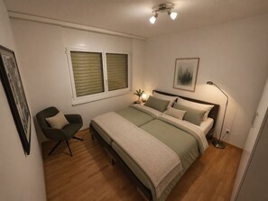 A bedroom with a bed, bedside table, lamp, chair, and a framed picture on the wall. - Ambassador A11 # 4-bed apartment, Dusche und Bad, WC by Interhome (Leukerbad)
