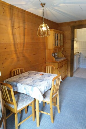 Een houten interieur met een eettafel, stoelen, een hanglamp en een keukenhoek.