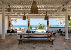 An outdoor patio with wooden tables and chairs, a view of the ocean, and hanging woven light fixtures.