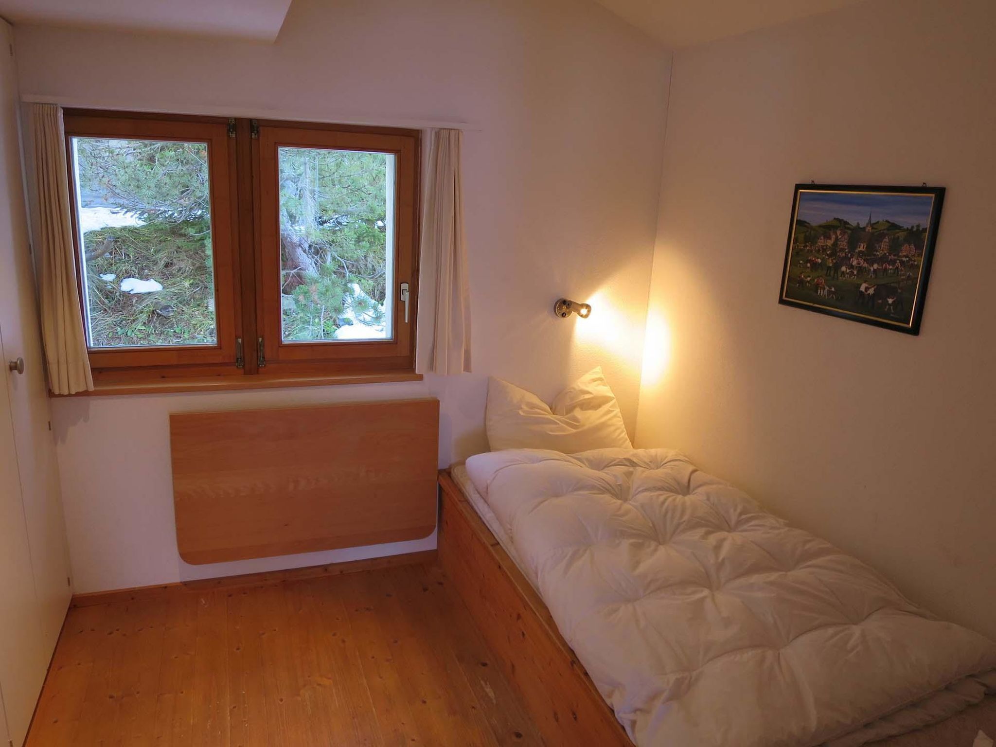 Ein kleines Zimmer mit einem Einzelbett, einem Holz-Kopfteil, einem Fenster mit Vorhängen, einer Wandleuchte und einem gerahmten Bild.