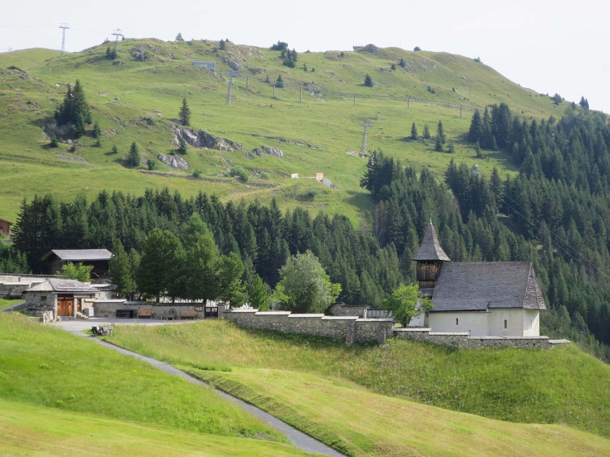 A rural setting with a stone building, a wooden structure with a pointed roof, and a pathway leading uphill.