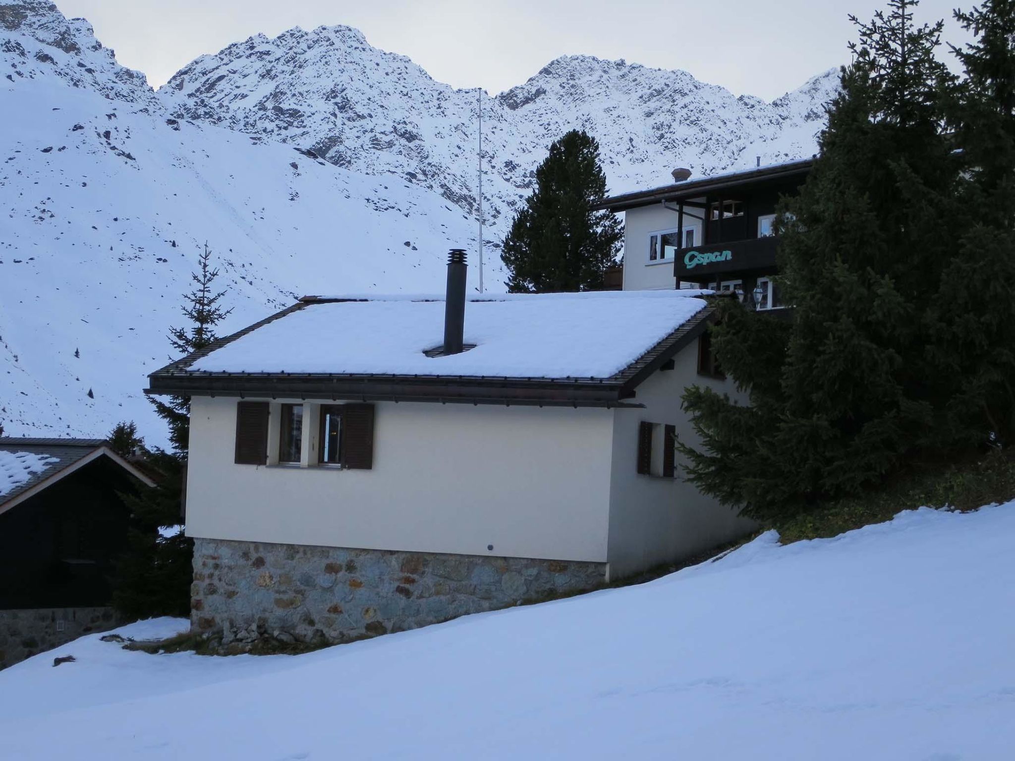 A snow-covered mountain house with a chimney, a stone foundation, and a view of snow-covered peaks.