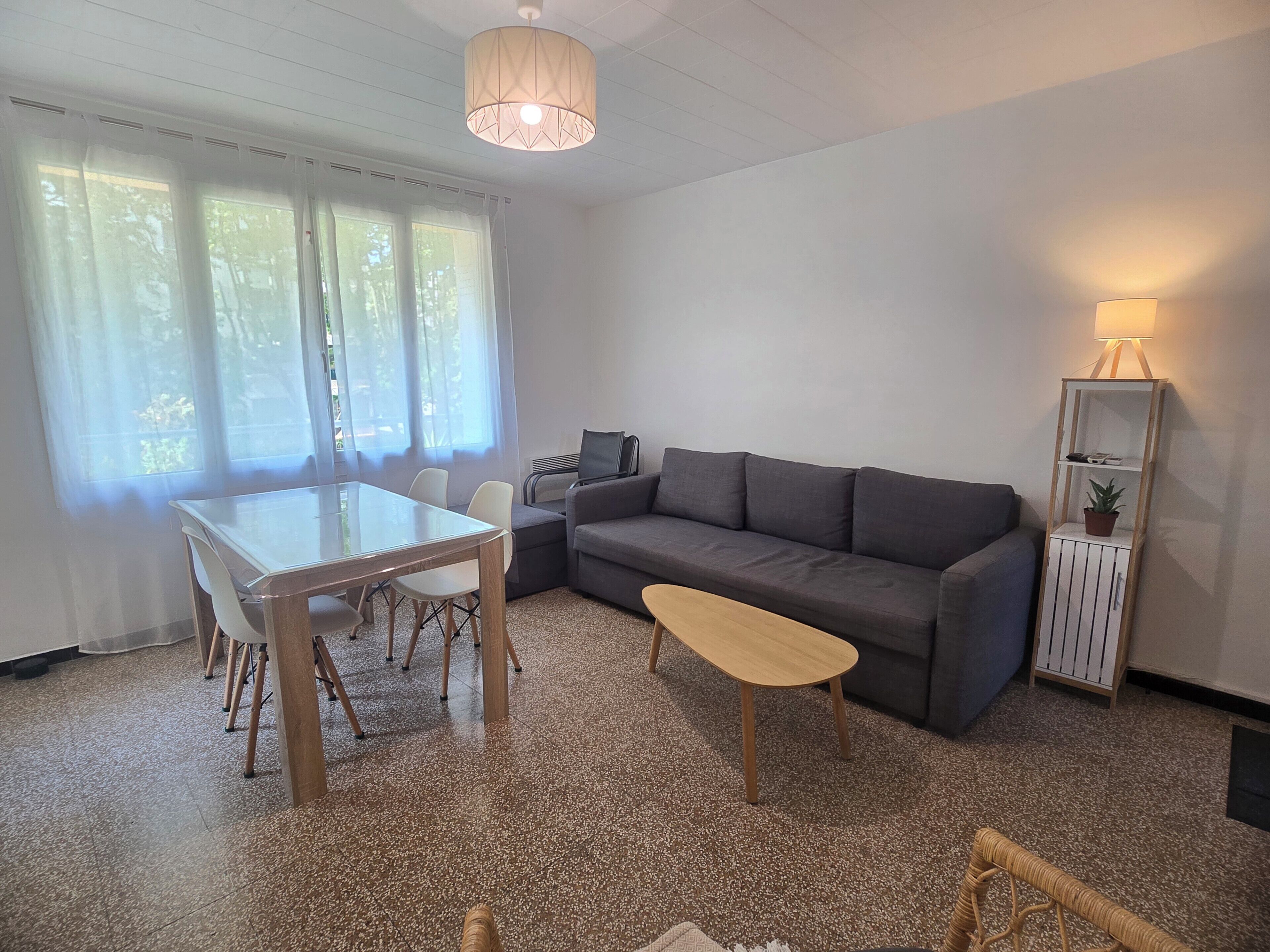 Ein Zimmer mit Glastisch, grauem Sofa, einer Holzbank, einer Stehlampe und einer kleinen Pflanze auf einem weißen Schrank.