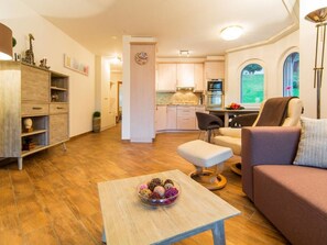 A modern living room with a sofa, a coffee table with a decorative bowl, a kitchen with cabinets and a window, and a hallway leading to another room. - Ambassador B13 # 4-bed apartment, Dusche und Bad, WC by Interhome (Leukerbad)