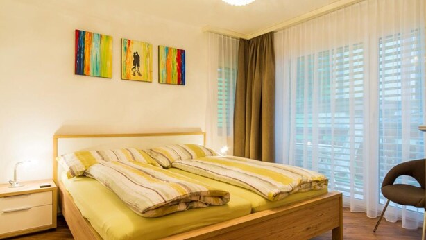 A bedroom with a wooden bed, a nightstand, a lamp, and a chair. There are three paintings on the wall. - Ambassador B13 # 4-bed apartment, Dusche und Bad, WC by Interhome (Leukerbad)