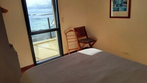 A room with a bed, a chair, a table, a window with a view of the sea, and a framed picture on the wall.