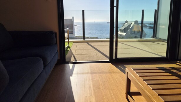 A modern living room with a sofa, a wooden bench, and a large sliding glass door leading to a balcony with a view of the sea.