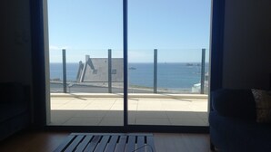 A room with a large window overlooking a coastal area, featuring a view of the sea, a lighthouse, and distant ships.