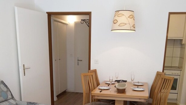 Um espaço de estar compacto com uma mesa de jantar posta para quatro pessoas, rodeada por cadeiras de madeira. Um sofá está posicionado à esquerda, e uma pequena mesa de madeira em primeiro plano.