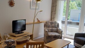 Uma sala de estar com televisão, uma mesa de jantar com cadeiras, um sofá, um abajur e uma janela com cortinas.